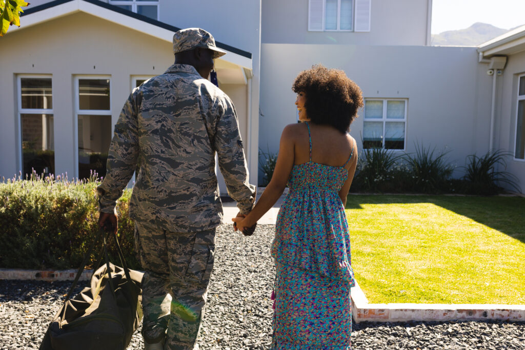 A multiracial military couple. Air Force service member walking in his camouflage uniform with his duffle bag. He is holding his girlfriend's hand and they are laughing, smiling, and walking into a house.