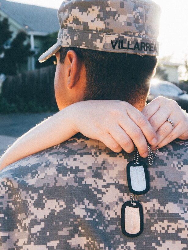 Army service member with his spouse. He is facing away and she is hugging him and holding dog tags.