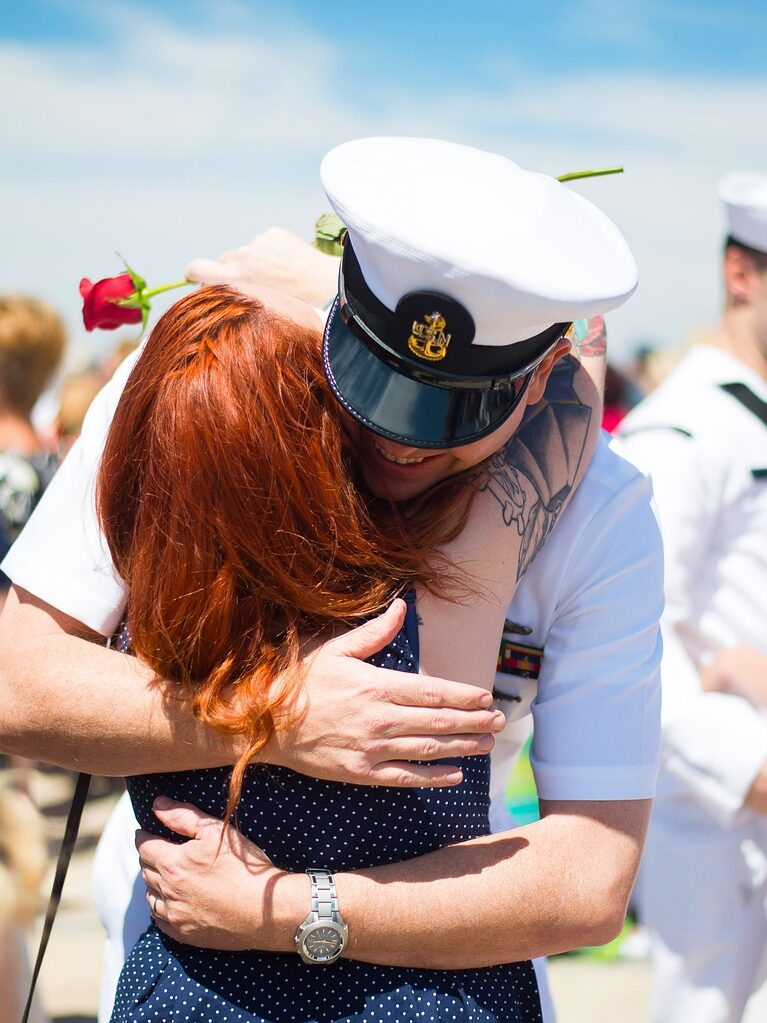 Navy service members in uniform at a homecoming. They have just come off the boat or submarine and are giving hugs to their family members and loved ones. One sailor is hugging his wife and she has a red rose in her hands.